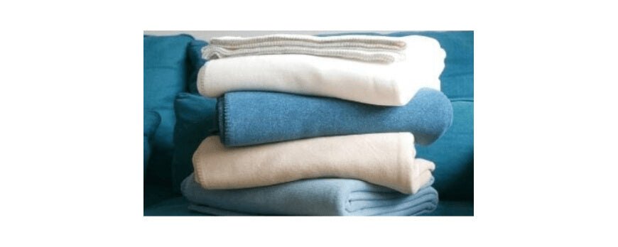 Pile de plaids polaires écru, bleu jean et rose poudré sur un canapé bleu – Plaid Addict
