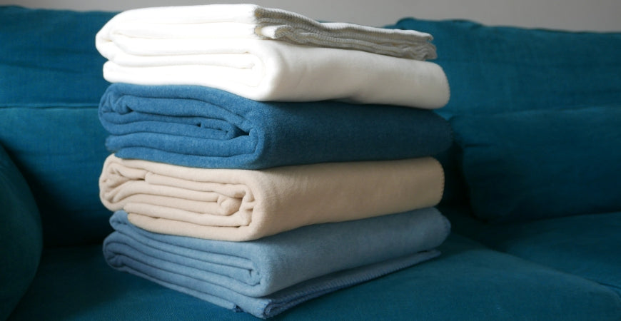 Pile de plaids polaires blanc, bleu et beige sur un canapé pétrole – Plaid Addict